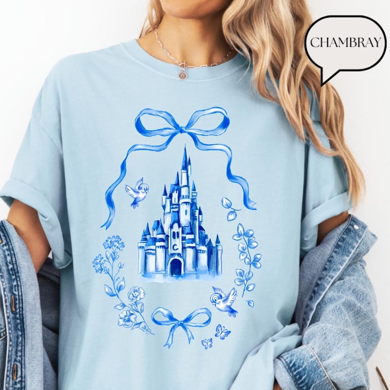 以下が含まれることがあります： 水色のTシャツ。水彩画のイラストで、お城、鳥、リボン、青色の花柄が描かれています。Tシャツには「CHAMBRAY」という言葉が吹き出しの中にあります。