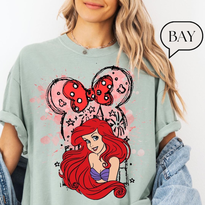 May include: A light green t-shirt featuring a graphic of Ariel from The Little Mermaid. The design includes a red bow, Mickey Mouse ears, and a pink watercolor splash. The word "BAY" is in a speech bubble.