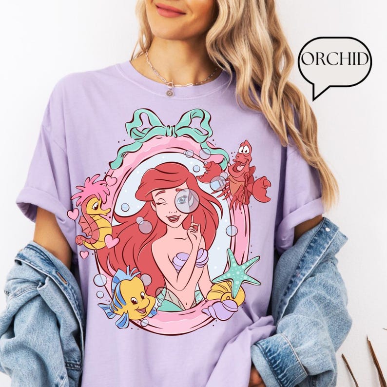 May include: A lilac-colored t-shirt featuring a cartoon illustration of Ariel from The Little Mermaid. The design includes Ariel winking with a magnifying glass, surrounded by sea creatures within a decorative frame. The word "ORCHID" is in a speech bubble.