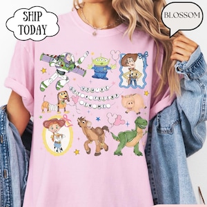 Puede incluir: Una camiseta de color rosa claro con un estampado gr&aacute;fico de varios personajes animados, incluyendo un ranger espacial, un vaquero, una vaquera, un dinosaurio verde, un cerdo, un perro y un alien&iacute;gena. El texto "YOU'VE GOT A FRIEND IN ME" est&aacute; impreso en el centro.