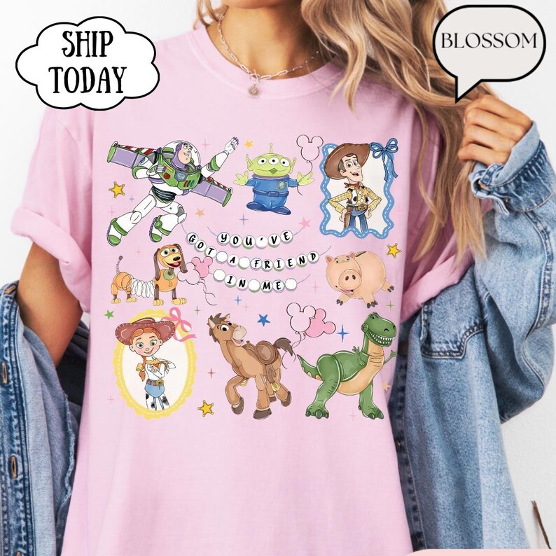 Puede incluir: Una camiseta de color rosa claro con un estampado gr&aacute;fico de varios personajes animados, incluyendo un ranger espacial, un vaquero, una vaquera, un dinosaurio verde, un cerdo, un perro y un alien&iacute;gena. El texto "YOU'VE GOT A FRIEND IN ME" est&aacute; impreso en el centro.