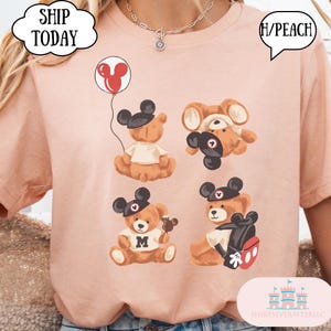 May include: A peach-colored t-shirt featuring four brown teddy bears wearing black mouse-ear hats. One bear holds a red balloon, one is upside down, one wears a shirt with the letter M, and one carries a black backpack with red accents.