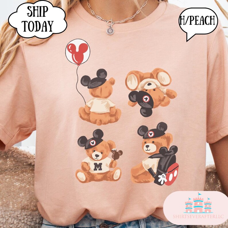 May include: A peach-colored t-shirt featuring four brown teddy bears wearing black mouse-ear hats. One bear holds a red balloon, one is upside down, one wears a shirt with the letter M, and one carries a black backpack with red accents.