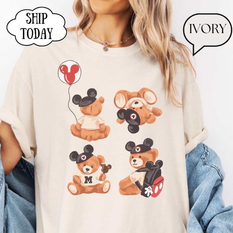 May include: An ivory t-shirt featuring a graphic print of four brown teddy bears wearing black mouse ear hats. One bear holds a red balloon, one is upside down, one holds a small treat, and one carries a black and red backpack. Text reads SHIP TODAY and IVORY.