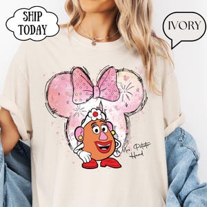 Può includere: Una t-shirt color avorio con una grafica di Mrs. Potato Head che indossa un fiocco rosa a pois, su uno sfondo a forma di orecchie di Topolino rosa in stile acquerello con fuochi d'artificio e il testo Mrs. Potato Head in corsivo nero.