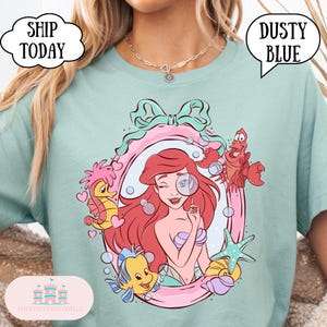 May include: Dusty blue t-shirt featuring a cartoon illustration of Ariel from The Little Mermaid, framed by a pink and teal design. The graphic includes seahorse, fish, and crab characters. Text on the shirt reads "SHIP TODAY".