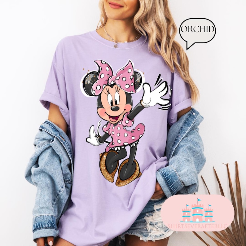 Puede incluir: Una camiseta morada con un gr&aacute;fico de Minnie Mouse con un vestido rosa de lunares y un lazo rojo. El texto "ORCHID" est&aacute; en un bocadillo en la esquina superior derecha de la imagen.