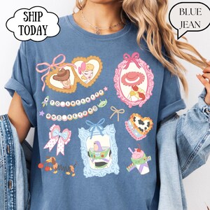 Op de afbeelding: Blauwe jeanskleurige t-shirt met kleurrijke cartoonkarakterontwerpen. Het shirt heeft de tekst "TO INFINITY AND BEYOND" en andere cartoonkarakterontwerpen. Het shirt heeft opgerolde mouwen en een relaxte pasvorm.