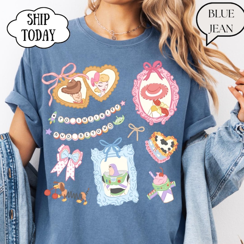 Op de afbeelding: Blauwe jeanskleurige t-shirt met kleurrijke cartoonkarakterontwerpen. Het shirt heeft de tekst "TO INFINITY AND BEYOND" en andere cartoonkarakterontwerpen. Het shirt heeft opgerolde mouwen en een relaxte pasvorm.