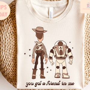 May include: A beige t-shirt featuring a brown cartoon image of Woody and Buzz Lightyear from Toy Story. The text "you got a friend in me" is printed below the characters.