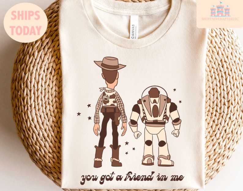 May include: A beige t-shirt featuring a brown cartoon image of Woody and Buzz Lightyear from Toy Story. The text "you got a friend in me" is printed below the characters.