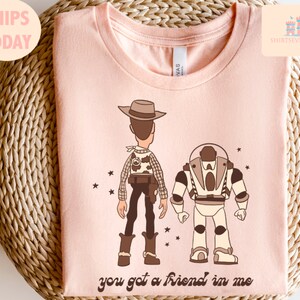 May include: Pink t-shirt featuring a cartoon image of Woody and Buzz Lightyear from Toy Story. The text "you got a friend in me" is printed below the characters.