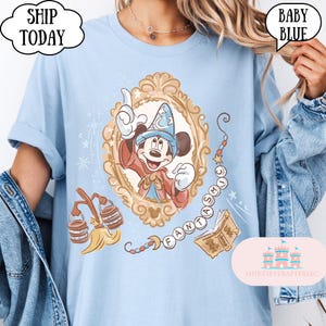 K&ouml;nnte beinhalten: Hellblaues T-Shirt mit einer gerahmten Illustration von Mickey Mouse als Zauberer. Das Design enth&auml;lt das Wort "FANTASMIC" und andere magische Elemente. Das Shirt hat den Text "SHIP TODAY" in einer wolkenf&ouml;rmigen Blase.