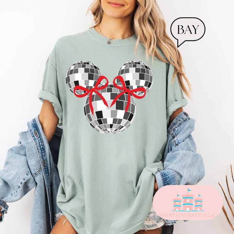 May include: A light green t-shirt featuring a black and white disco ball design in the shape of Mickey Mouse ears. The disco balls have red bows on them.