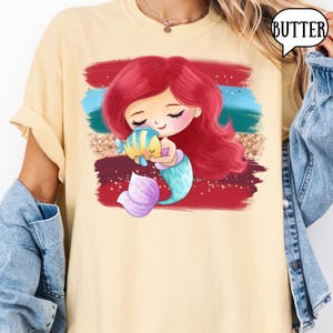 Puede incluir: Camiseta amarillo claro con un dise&ntilde;o de sirena de dibujos animados. La sirena tiene el pelo rojo y una cola turquesa, sosteniendo un pez amarillo y azul. El gr&aacute;fico est&aacute; sobre pinceladas rojas, turquesas y doradas. La palabra "BUTTER" est&aacute; en un bocadillo.