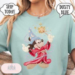 May include: A light blue t-shirt with a cartoon Mickey Mouse as a wizard, holding a wand with stars. The design includes a blue wizard hat and a red robe. The shirt has the text "Dusty Blue" in a speech bubble.