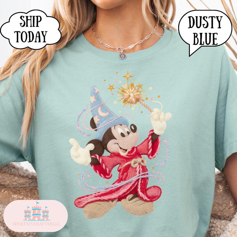 May include: A light blue t-shirt with a cartoon Mickey Mouse as a wizard, holding a wand with stars. The design includes a blue wizard hat and a red robe. The shirt has the text "Dusty Blue" in a speech bubble.