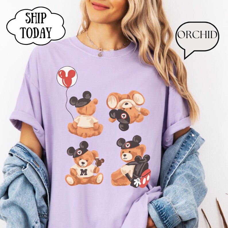 May include: A light purple t-shirt featuring a graphic of four brown teddy bears wearing black mouse ear hats. One bear holds a red balloon, another wears a white shirt with an M, and one carries a black backpack. Text includes SHIP TODAY and ORCHID.