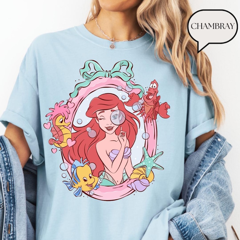 May include: Light blue t-shirt featuring a cartoon illustration of Ariel from The Little Mermaid. The design includes Ariel, Flounder, Sebastian, and a seahorse within a pink and green frame. The word "CHAMBRAY" is in a speech bubble.