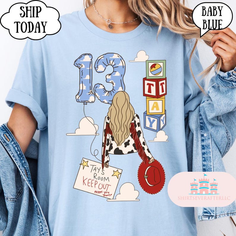 May include: A light blue t-shirt featuring a graphic of a person with long blonde hair wearing a cow-print jacket with red fringe. The design includes a balloon shaped like the number 13, stacked alphabet blocks, and a sign reading Tay's Room Keep Out.