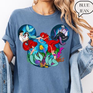 Puede incluir: Camiseta azul vaquero con un gr&aacute;fico de Ariel y otros personajes de La Sirenita. El dise&ntilde;o tiene forma de orejas de Mickey Mouse. La camiseta es de estilo c&oacute;modo e informal.