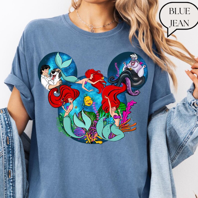 Puede incluir: Camiseta azul vaquero con un gr&aacute;fico de Ariel y otros personajes de La Sirenita. El dise&ntilde;o tiene forma de orejas de Mickey Mouse. La camiseta es de estilo c&oacute;modo e informal.