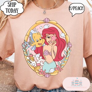 May include: Peach-colored t-shirt featuring a cartoon illustration of Ariel from The Little Mermaid, holding a starfish with Flounder. The design is framed in a gold oval with seashells and floral accents. The shirt has the text "H/PEACH".