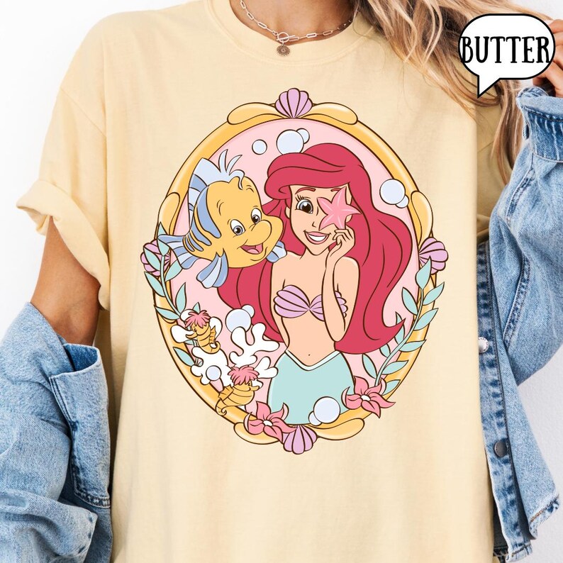May include: A light yellow t-shirt featuring a cartoon illustration of Ariel from The Little Mermaid. Ariel has red hair and is holding a pink starfish. Flounder the fish is next to her. The word "BUTTER" is in a speech bubble.