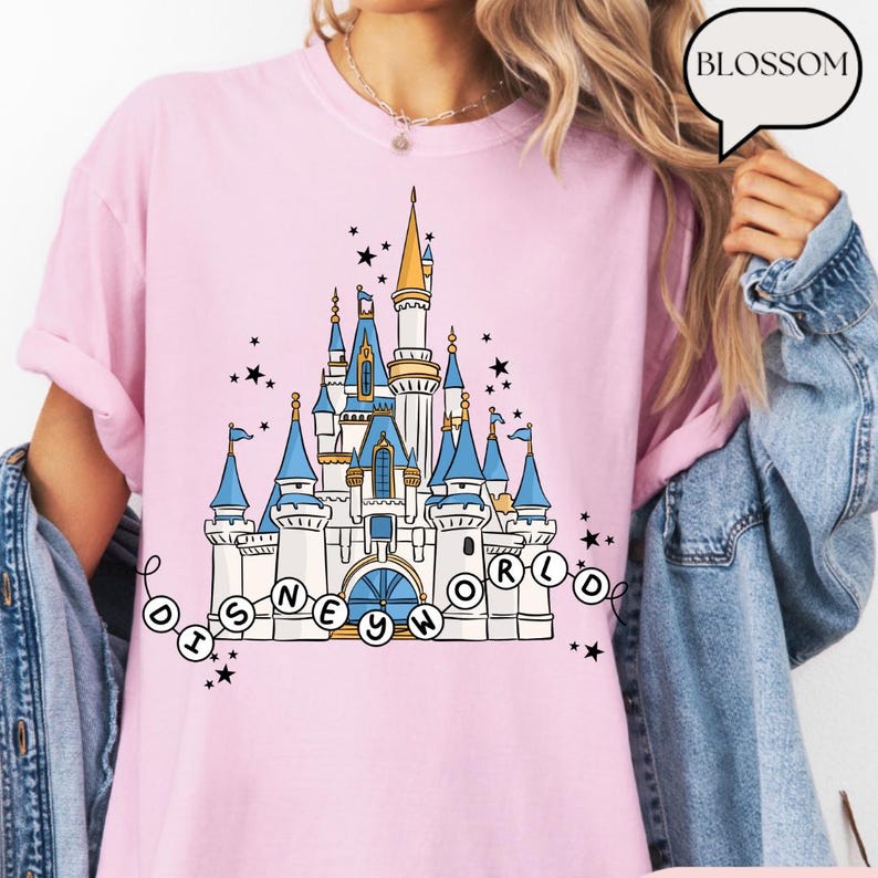 May include: Pink t-shirt featuring a detailed illustration of a castle with blue turrets and gold accents, with the text "DISNEY WORLD" in a circular design. Black stars are scattered around the castle. The word "BLOSSOM" is in a speech bubble.