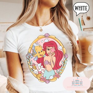 May include: White t-shirt featuring a colorful illustration of Ariel from The Little Mermaid, holding a starfish with Flounder. The design is framed in a gold-colored oval with a pink background and floral accents. The word "WHITE" is in a speech bubble.