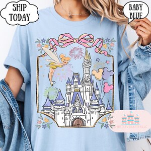 Puede incluir: Camiseta azul claro con una ilustraci&oacute;n de un castillo de cuento de hadas, un hada, fuegos artificiales y globos de Mickey Mouse. El dise&ntilde;o incluye un lazo rosa y detalles florales. El texto en la camiseta dice "SHIP TODAY" y "BABY BLUE".