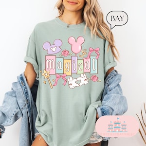 Op de afbeelding: Saliegroen t-shirt met het woord "magical" in pastelkleuren, met cartoonafbeeldingen van een toverstaf, Mickey Mouse-oren, een ballon en een konijn. Het shirt heeft opgerolde mouwen en een relaxte pasvorm.