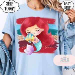 Puede incluir: Camiseta azul beb&eacute; con un dise&ntilde;o de sirena de dibujos animados. La sirena tiene el pelo rojo, una cola turquesa y sostiene un pez amarillo y azul. El fondo presenta pinceladas rojas, turquesas y doradas. El texto en la camiseta dice "Baby Blue" y "Ship Today".
