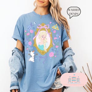 K&ouml;nnte beinhalten: Ein hellblaues T-Shirt mit einem Aufdruck von Elsa aus Frozen. Der Aufdruck zeigt Elsa in einem goldenen Rahmen mit Blumen und einer Schleife. Der Text "Washed Denim" ist auf dem Bild.
