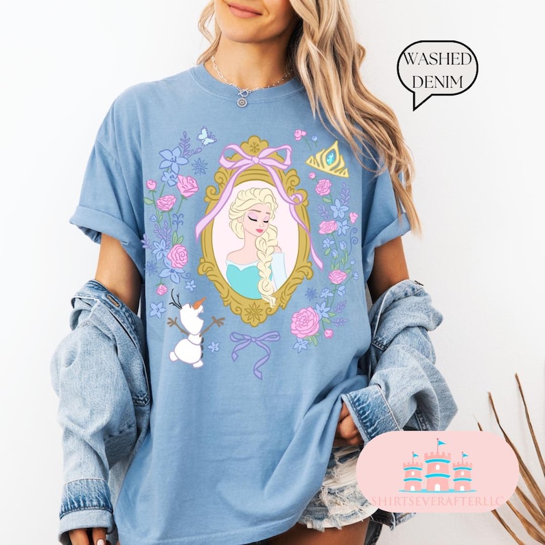 K&ouml;nnte beinhalten: Ein hellblaues T-Shirt mit einem Aufdruck von Elsa aus Frozen. Der Aufdruck zeigt Elsa in einem goldenen Rahmen mit Blumen und einer Schleife. Der Text "Washed Denim" ist auf dem Bild.