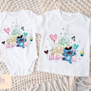 May include: Two white t-shirts with a colorful illustration of a cartoon character in front of a castle. The character is wearing Mickey Mouse ears and holding a balloon. The castle is surrounded by colorful hearts and Mickey Mouse ears.