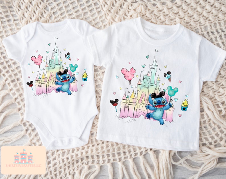 May include: Two white t-shirts with a colorful illustration of a cartoon character in front of a castle. The character is wearing Mickey Mouse ears and holding a balloon. The castle is surrounded by colorful hearts and Mickey Mouse ears.