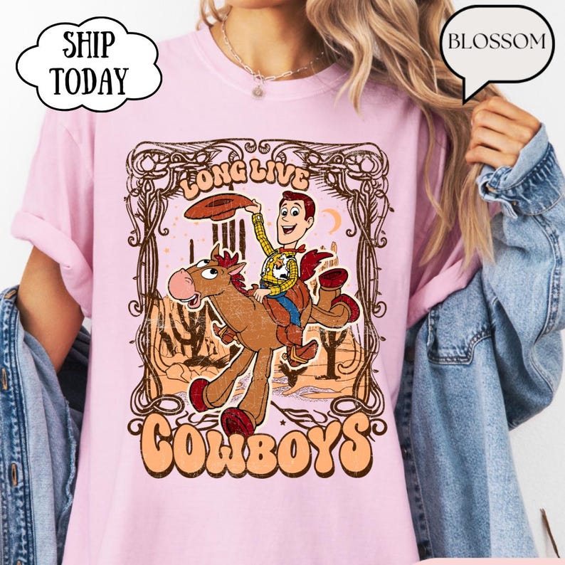 May include: Pink t-shirt featuring a cartoon cowboy riding a horse, with the words "Long Live Cowboys" in a vintage style. The design includes a desert landscape and the cowboy is wearing a hat. The shirt is a light pink color.