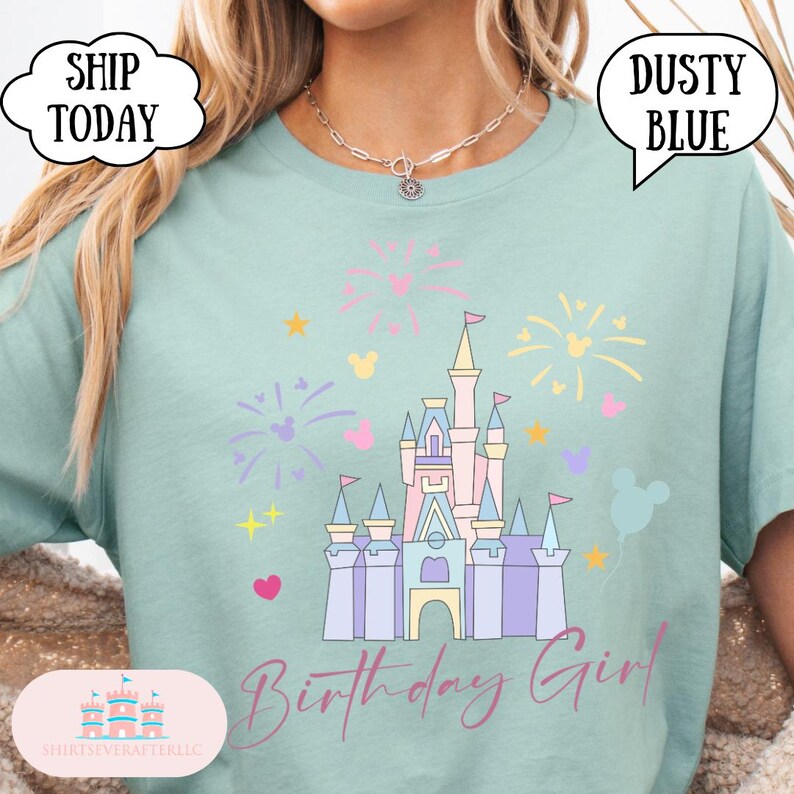 May include: A pale blue t-shirt with a castle graphic, fireworks, and Mickey Mouse shapes. The words "Birthday Girl" are in a script font. The shirt has a loose fit. The text "Dusty Blue" is in a speech bubble.