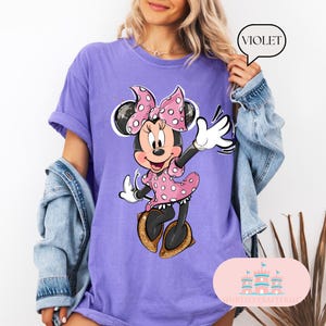 Puede incluir: Camiseta morada con un gr&aacute;fico de Minnie Mouse con un vestido rosa de lunares y un lazo rojo. El texto "VIOLET" est&aacute; en un bocadillo en la esquina superior derecha de la imagen.