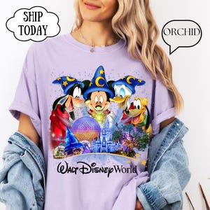 May include: A light purple t-shirt featuring Mickey Mouse, Goofy, Donald Duck, and Pluto in wizard hats, with a Walt Disney World graphic. The shirt has rolled-up sleeves and is paired with a denim jacket. The text "SHIP TODAY" is in a white bubble.