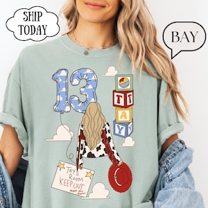 May include: A sage green t-shirt featuring a graphic of a person with long blonde hair wearing a cow-print jacket with red fringe. The design includes a blue cloud-patterned number 13 balloon, alphabet blocks, and a sign reading Tays Room Keep Out.