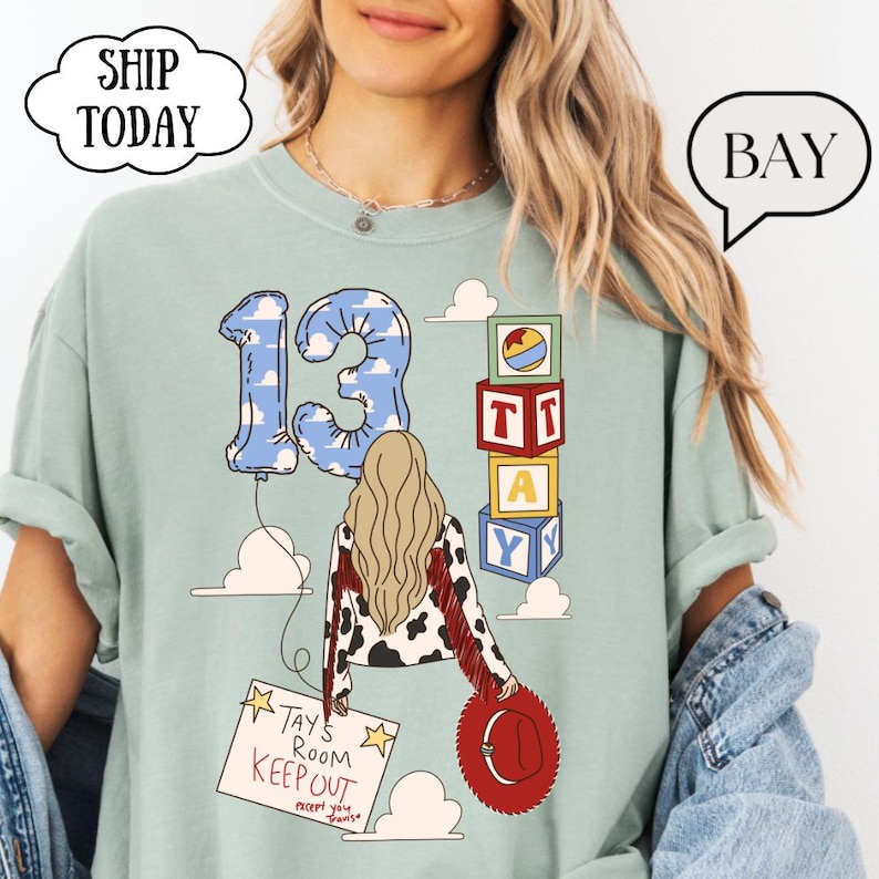 May include: A sage green t-shirt featuring a graphic of a person with long blonde hair wearing a cow-print jacket with red fringe. The design includes a blue cloud-patterned number 13 balloon, alphabet blocks, and a sign reading Tays Room Keep Out.