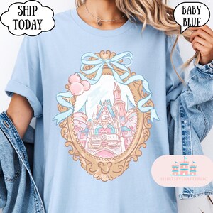 Puede incluir: Camiseta azul beb&eacute; con una ilustraci&oacute;n enmarcada en oro de un castillo de cuento de hadas con detalles en rosa y azul. El dise&ntilde;o incluye un lazo y globos. El texto en la camiseta dice "BABY BLUE" y "SHIP TODAY".
