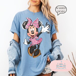 Puede incluir: Una camiseta azul claro con un gr&aacute;fico de Minnie Mouse con un vestido rosa de lunares y un lazo rosa. El texto "WASHED DENIM" est&aacute; en un cuadro blanco en la esquina superior derecha de la imagen.