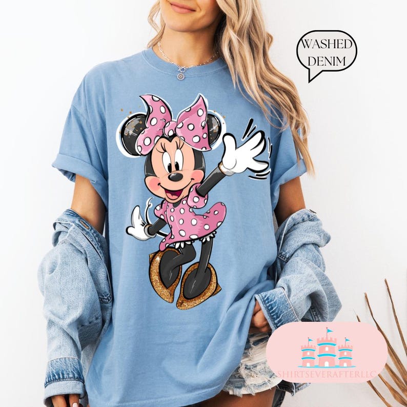 Puede incluir: Una camiseta azul claro con un gr&aacute;fico de Minnie Mouse con un vestido rosa de lunares y un lazo rosa. El texto "WASHED DENIM" est&aacute; en un cuadro blanco en la esquina superior derecha de la imagen.