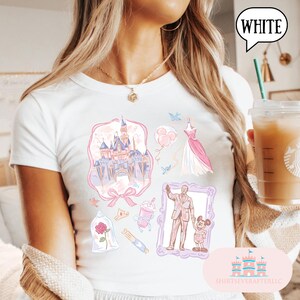 Op de afbeelding: Wit T-shirt met een kleurrijk grafisch ontwerp met een kasteel, jurk, ballonnen en andere illustraties. Het shirt heeft het woord "WHITE" in een tekstballon. Het shirt heeft ook de tekst "SHIRTSEVERAFTERLLC".