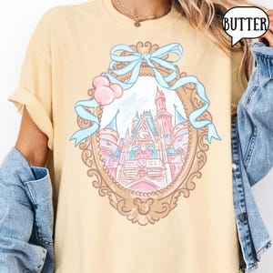 Puede incluir: Camiseta amarilla clara con una ilustraci&oacute;n de un castillo de cuento de hadas dentro de un marco ornamentado, adornada con una cinta azul claro y globos rosas. La palabra "BUTTER" est&aacute; en una burbuja. El dise&ntilde;o est&aacute; centrado en la camiseta.