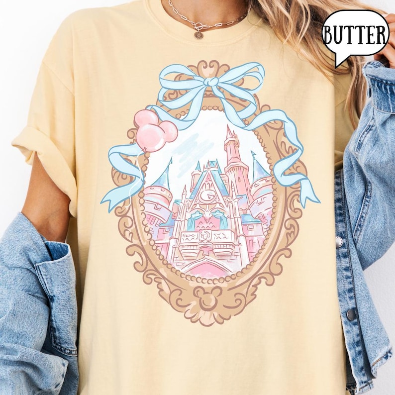 Puede incluir: Camiseta amarilla clara con una ilustraci&oacute;n de un castillo de cuento de hadas dentro de un marco ornamentado, adornada con una cinta azul claro y globos rosas. La palabra "BUTTER" est&aacute; en una burbuja. El dise&ntilde;o est&aacute; centrado en la camiseta.