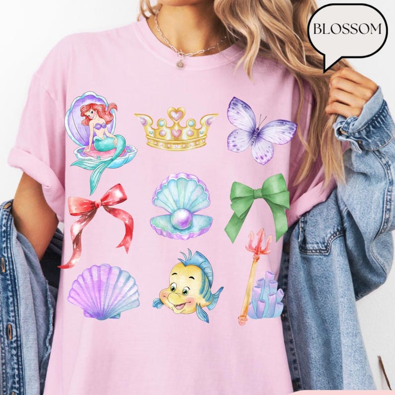 Może przedstawiać: R&oacute;żowa koszulka z akwarelowymi ilustracjami syreny, korony, motyla, kokardy, muszli i Florka. Koszulka ma r&oacute;wnież słowo "BLOSSOM" w dymku.